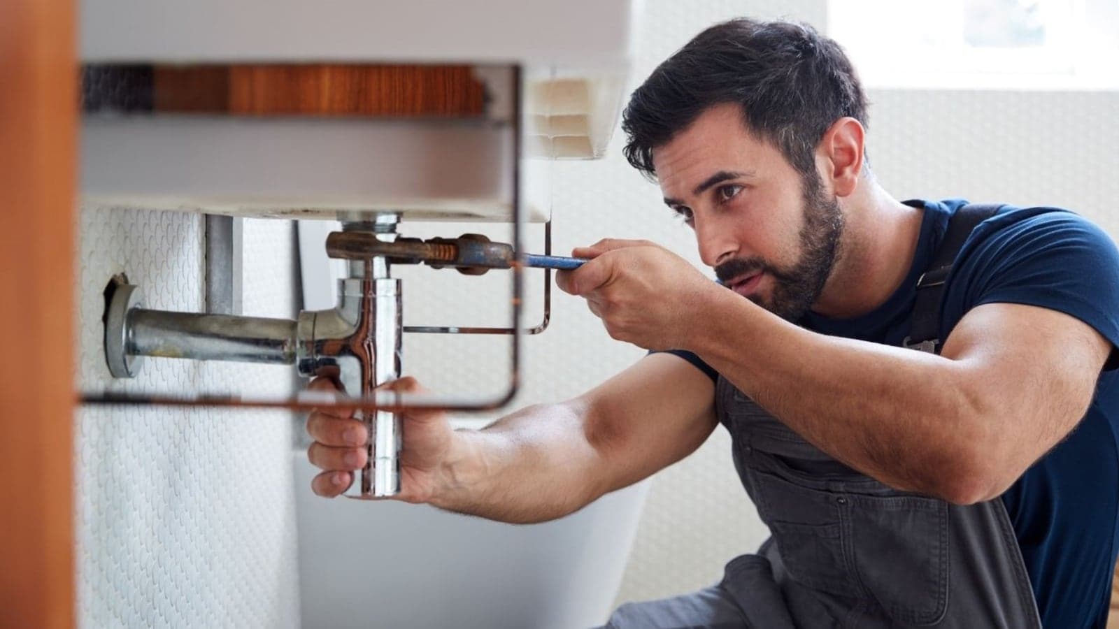 A plumber using a wrench on the waste trap and pipes under a sink.